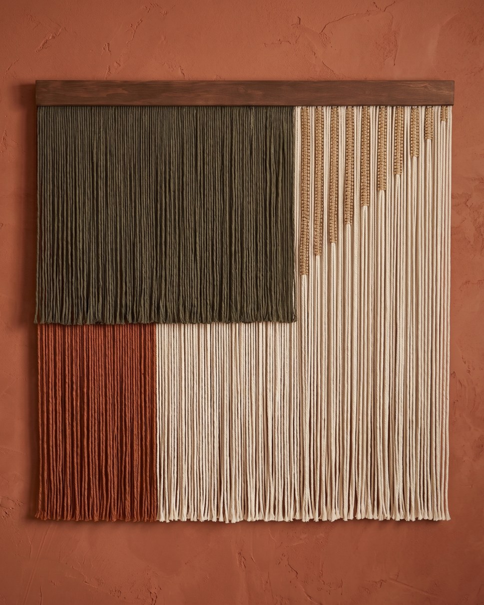 A handwoven macrame wall hanging with vertical panels of forest green, terracotta, and cream cotton, accented with gold woven details, hanging from a dark wooden dowel against a terracotta wall.